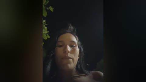 Video of a young woman with long black hair, dark skin, and a serious expression, partially obscured by a shadowy, indistinct background. She has a bandage on her nose and is wearing a dark top.