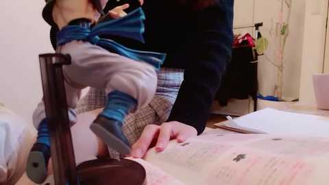 Media: Video of a person in a blue jacket and plaid shorts, leaning over a desk with a book, in a cluttered room with medical equipment.