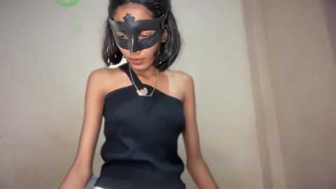 Video of a slim, East Asian woman with straight black hair, wearing a black leather mask and sleeveless black top, standing in a beige-walled room.
