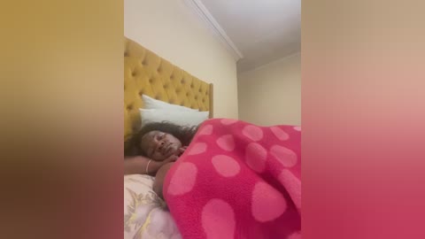 A video of a woman with light skin, curly hair, and a red polka-dot blanket covering her, lying in a bed with a yellow upholstered headboard and light-colored bedding.