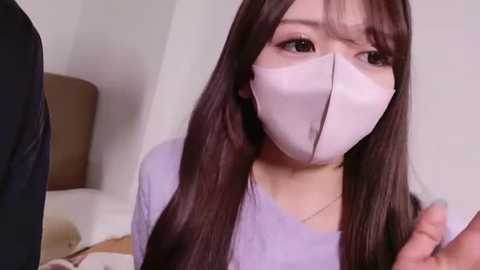 Video of an East Asian woman with long black hair, wearing a white face mask, light lavender shirt, and a necklace. She is indoors, in a simple room with a beige lamp and a white wall.