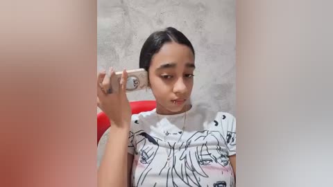 Video of a young girl with medium skin tone, black hair, and a white t-shirt featuring black line drawings of faces. She is using a beige flip phone to her ear, sitting in front of a textured gray wall.