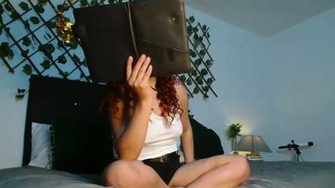 Video of a woman with curly red hair, wearing a white tank top and black shorts, sitting on a bed, covering her face with her hands, in a minimalist bedroom with a dark headboard and green wall decor.