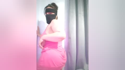 A video of a woman in a pink dress, black face mask, and headscarf, standing in a hallway with light pink and gray curtains.