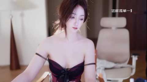 Video of a young, light-skinned East Asian woman with shoulder-length brown hair, wearing a black and burgundy lace bra, sitting on a beige office chair in a softly lit room with wooden floors.