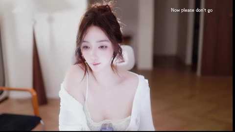 A video of a young woman with pale skin and dark hair in an off-shoulder white top, sitting in a modern, dimly lit room with wooden floors. Text reads, \"Now please don't go.\