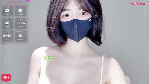 Video of an East Asian woman with straight black hair, wearing a black face mask and a white spaghetti-strap top, standing against a plain white background.