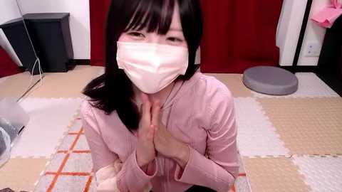 Video of an Asian woman with long black hair, wearing a pink cardigan, white face mask, and kneeling on a tatami mat. She's praying with hands clasped. Red curtains, a gray cushion, and a pink item are visible in the background.