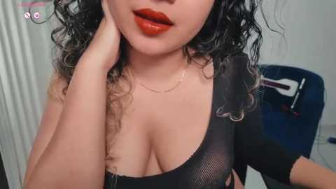 Video of a woman with light skin, curly black hair, and full red lips, wearing a black mesh top revealing cleavage, in a dimly lit room with a blue chair and white wall.