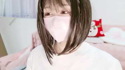 Video of an Asian woman with shoulder-length, straight black hair, wearing a pink surgical mask and a white shirt. Background shows a bed with pink sheets and a red pillow with white flowers.