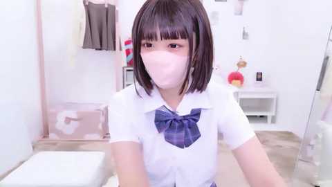 A young Asian girl with straight, dark hair and bangs, wearing a white shirt and blue plaid bow tie, sits in a bright, clean room with a white wall, shelves, and a red apple.