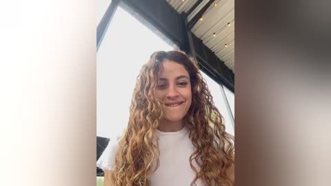 A video of a smiling woman with curly, light brown hair, wearing a white t-shirt, standing in front of large windows with a modern, industrial-style interior.