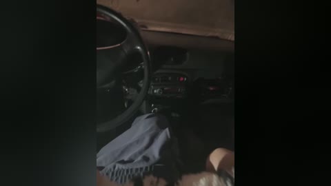 A dimly lit video of a car interior from the driver's perspective. The steering wheel and dashboard are visible, with the driver's hands on the wheel. The background is dark and indistinct, suggesting nighttime or a dimly lit environment.