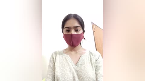 Video of a young woman with medium skin tone, dark hair, and brown eyes, wearing a red face mask and a beige patterned long-sleeve top. The background is a light-colored wall.
