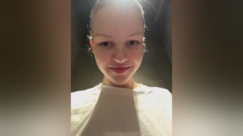 Video of a young woman with fair skin and dark hair tied back, smiling softly. She wears a white top and stands against a blurred, dark background.