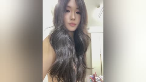 Video of a young Asian woman with long, wavy black hair, fair skin, and delicate features, standing in a modern bathroom with white tiles and a mirror. She is partially nude, wearing a neutral expression.