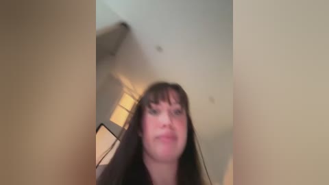 A blurred video of a young woman with long, dark hair and a neutral expression, taken indoors with beige walls and a ceiling fan visible.