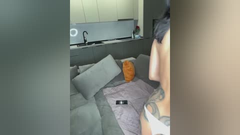 Media: Video of a modern, minimalist living room with a grey sectional sofa, a purple throw, and a large orange pillow. The background features a sleek kitchen with a black sink, a light-colored backsplash, and a round light fixture.