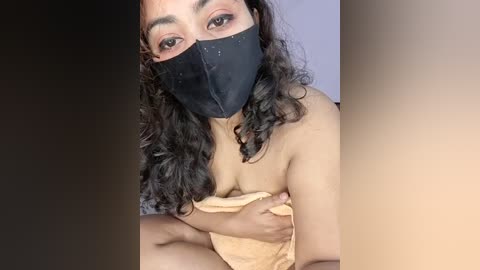 Video of a woman with medium brown skin, curly black hair, wearing a black mask and beige towel, covering her breasts, seated on a bed with a white pillow.
