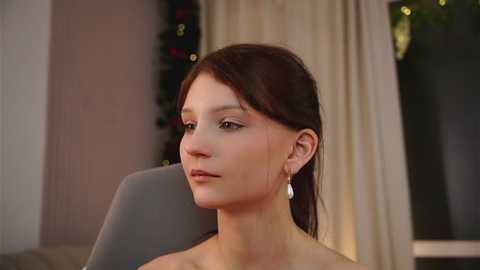 Video of a young Caucasian woman with fair skin and dark brown hair, wearing a simple dress, sitting indoors. She has a neutral expression and is looking to the left.