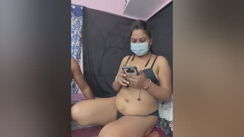 Media: A video of a plus-sized woman in a black bikini, wearing a mask, taking a selfie in a dimly lit room with colorful curtains.