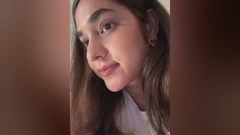 Media: A close-up video of a young woman with long, straight brown hair, light skin, and brown eyes, wearing a white top, smiling softly. The background is a blurred, warm-toned gradient.