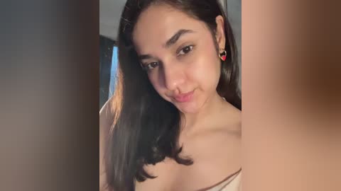 Video of a young woman with long dark hair, light skin, and a small heart earring, smiling softly, wearing a white off-shoulder top.