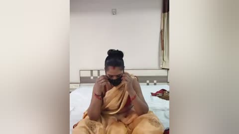 A video of a woman with medium brown skin and black hair in a bun, wearing a beige saree and black mask, seated on a bed in a simple, sparsely decorated room.
