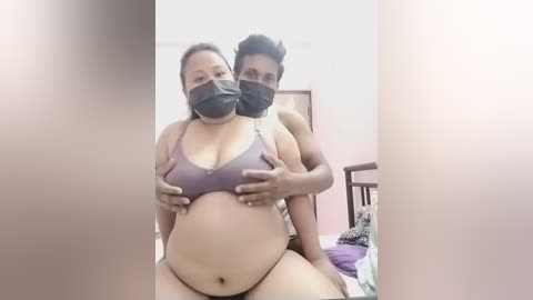 A video shows two pregnant women in a bedroom, wearing face masks. One woman sits on a bed, wearing a purple bra, and the other stands behind her, supporting her.