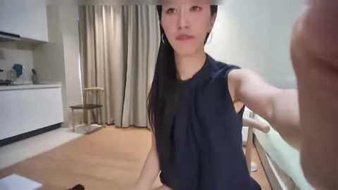 A video of an Asian woman with long black hair, wearing a dark sleeveless top, sitting on a wooden floor in a modern, minimalist room with white walls, beige curtains, and a white chair.