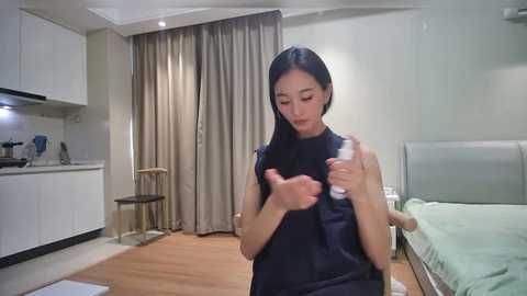Video of a petite, dark-haired Asian woman in a black sleeveless dress, holding a small, white object, standing in a modern, minimalist bedroom with light wood flooring, white cabinets, and a bed with light green sheets.