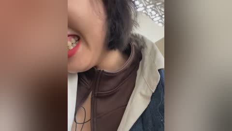 A close-up video of a woman with medium brown skin, smiling with visible braces, wearing a brown shirt and a denim jacket. Background features a beige wall and a grid ceiling.