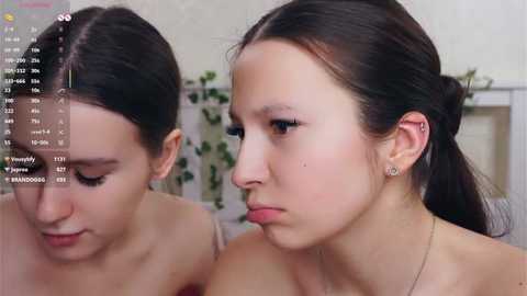 Video of two young women with light skin, dark hair, and minimal makeup, seated close, wearing strapless tops. The background shows a blurred indoor setting with a white wall and a potted plant.