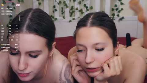 Video of two young, pale-skinned women with dark hair, topless, lying side by side, smiling, in a cozy room with green ivy and a red couch.