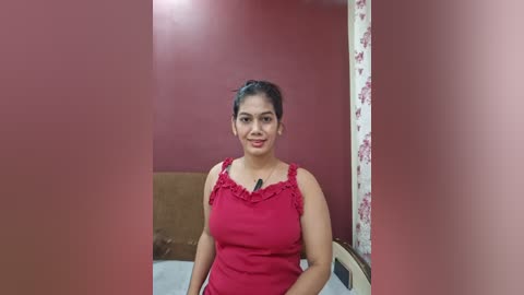 Video of a middle-aged woman with medium build, wearing a red sleeveless top, sitting on a bed in a room with red walls and floral-patterned curtains.