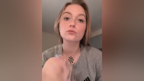 Media: Video of a young Caucasian woman with fair skin and short, light brown hair, wearing a gray shirt, blowing air onto a white object. Background shows a bathroom with white walls and a blue cabinet.