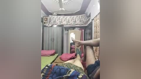 Video of a small, cluttered bedroom with a ceiling fan, floral curtains, and multiple beds covered in colorful blankets. A person is seen stretching their legs while wearing shorts.