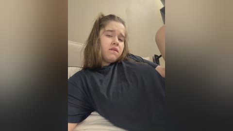 Video of a young Caucasian woman with long brown hair, lying on a bed in a dimly lit room, wearing a dark T-shirt, with blurred background.