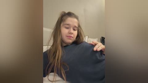 Video of a young girl with long brown hair, wearing a dark blue sweatshirt, lying on a bed with beige walls.