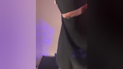 A video captures a close-up of a person wearing a black dress, standing in a dimly lit room with purple lighting. The focus is on the person's lower body and right hand, which is adorned with a ring.
