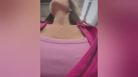 Video of a woman in a pink ribbed tank top, wearing a pink jacket, leaning forward with long brown hair. Background is blurred.