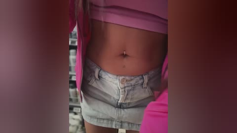 Video of a woman's midriff, wearing a pink shirt and light blue denim shorts, with a navel piercing and a small tattoo visible. Background shows a blurry store interior.