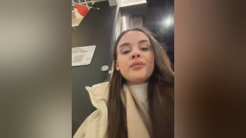 Video of a young woman with straight, long brown hair, wearing a beige jacket over a white turtleneck, standing in a dimly lit room with a black door and white signs on the wall.