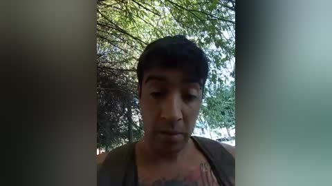 A video of a young man with short black hair, wearing a gray tank top, standing outdoors with a blurred background of green trees and sky. His facial expression is neutral.