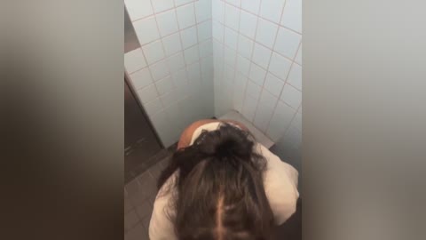 A video of a woman with long, dark hair tied in a bun, sitting on a toilet in a small, tiled bathroom with light blue walls and white tiles.