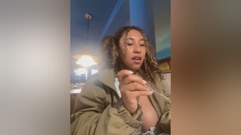 Video of a young Black woman with curly hair, wearing an open beige jacket, sitting indoors, looking contemplative. Background shows a dimly lit room with a ceiling fan and blue walls.
