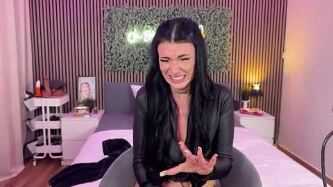 A video of a smiling woman with long black hair, wearing a black leather jacket, sitting on a bed in a modern bedroom with striped wallpaper, a green leaf wall, and a white bedside table with a lamp.