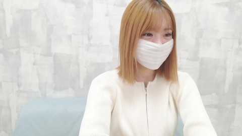 Video of an Asian woman with straight, shoulder-length, light brown hair, wearing a white mask and a beige jacket, seated in front of a textured, abstract background.