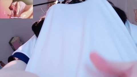 A video of a person wearing a white T-shirt with blue sleeves, blurred background showing a red object and a pink wall, with a small inset image of a person wearing a black shirt.