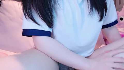 Media: Video of a young Asian woman with pale skin, wearing a white t-shirt with blue trim, sitting on a bed with pink sheets. Her long black hair falls over her shoulders.